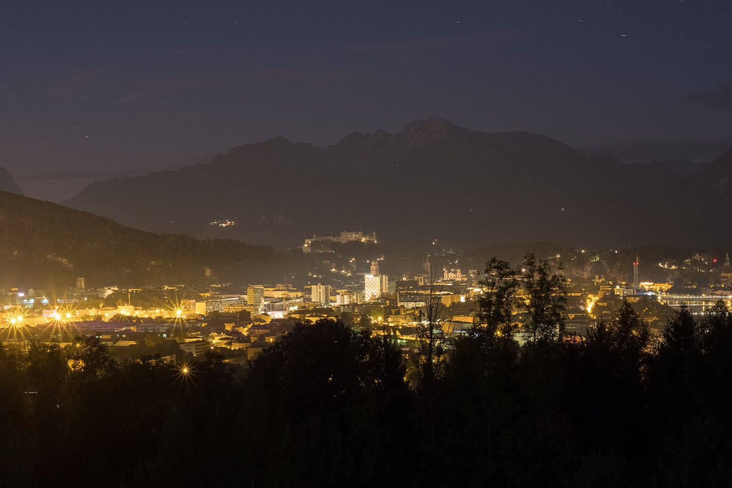 Salzburg city skyline lit up at night viewed from Maria Plain hill with Alps silhouette.