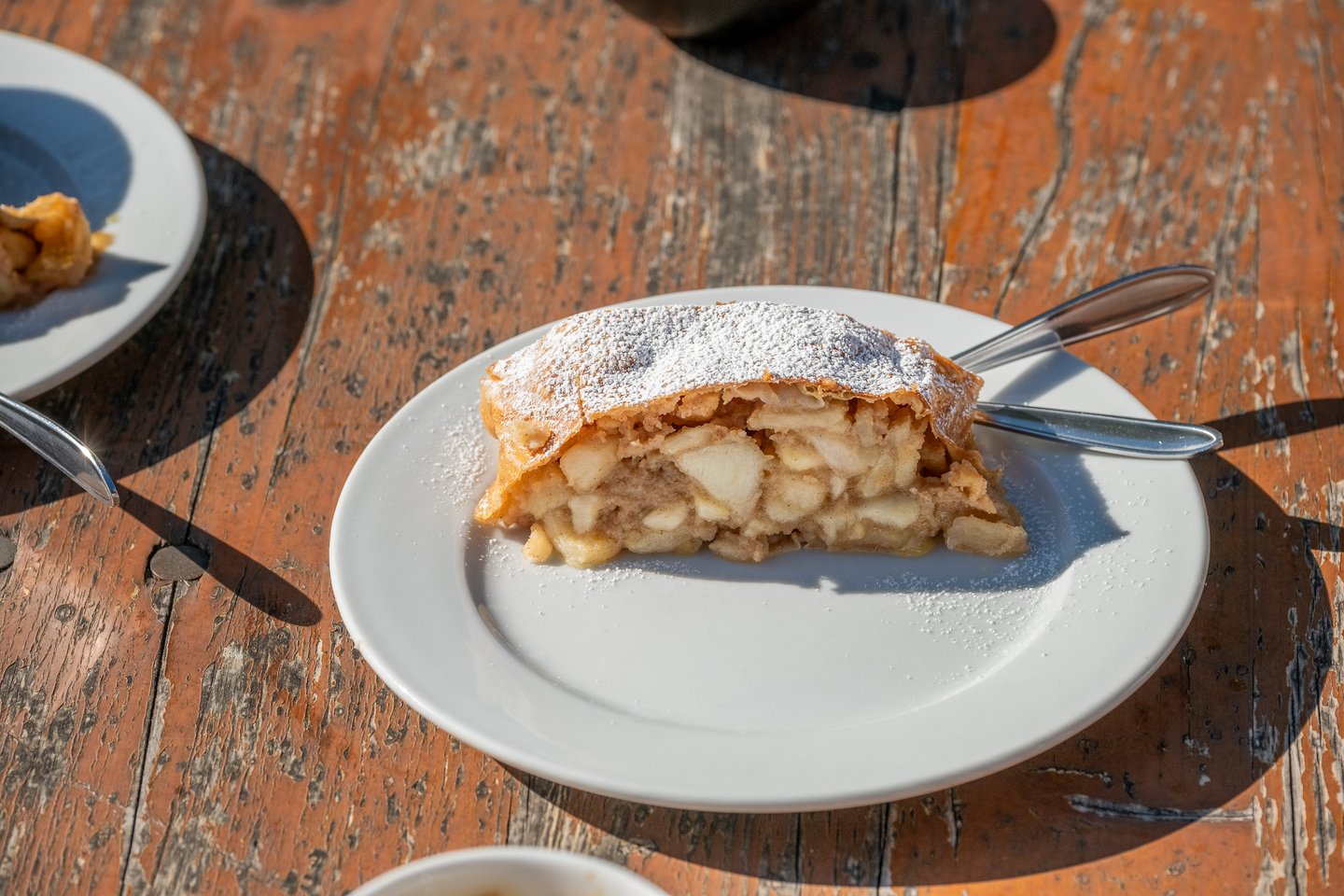 A slice of apple strudel dusted with icing sugar sits on a white plate on a weathered wooden table in bright sun at Werfen.