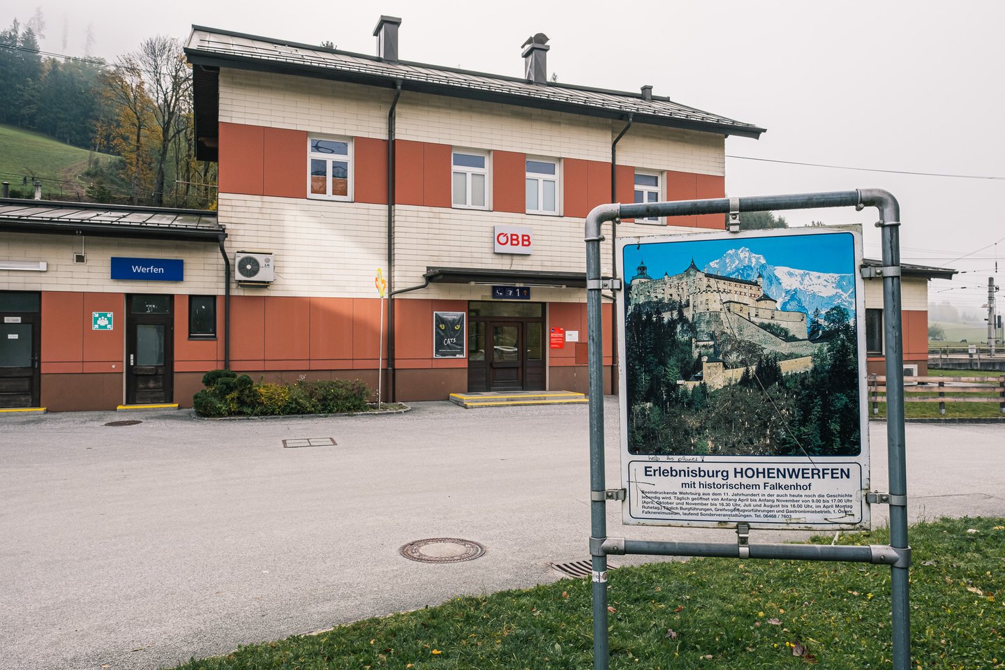 The cream and red Werfen station building seen from the forecourt, with the OeBB logo above the entrance and a weathered Hohenwerfen fortress billboard standing on the grass to the right.