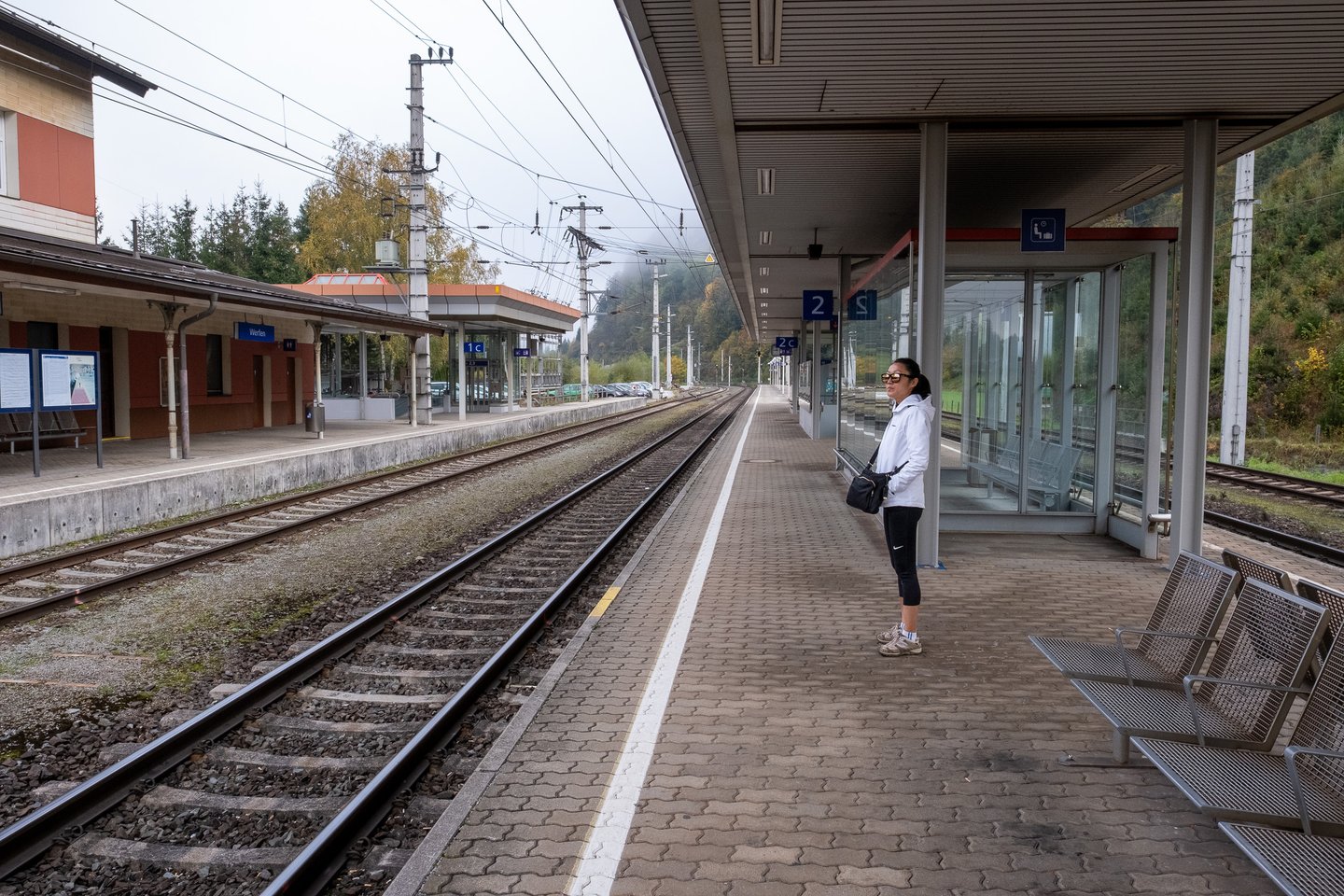 A woman in a white jacket waits alone on platform 2 at Werfen station on a misty autumn morning, with empty tracks running away between wooded slopes.