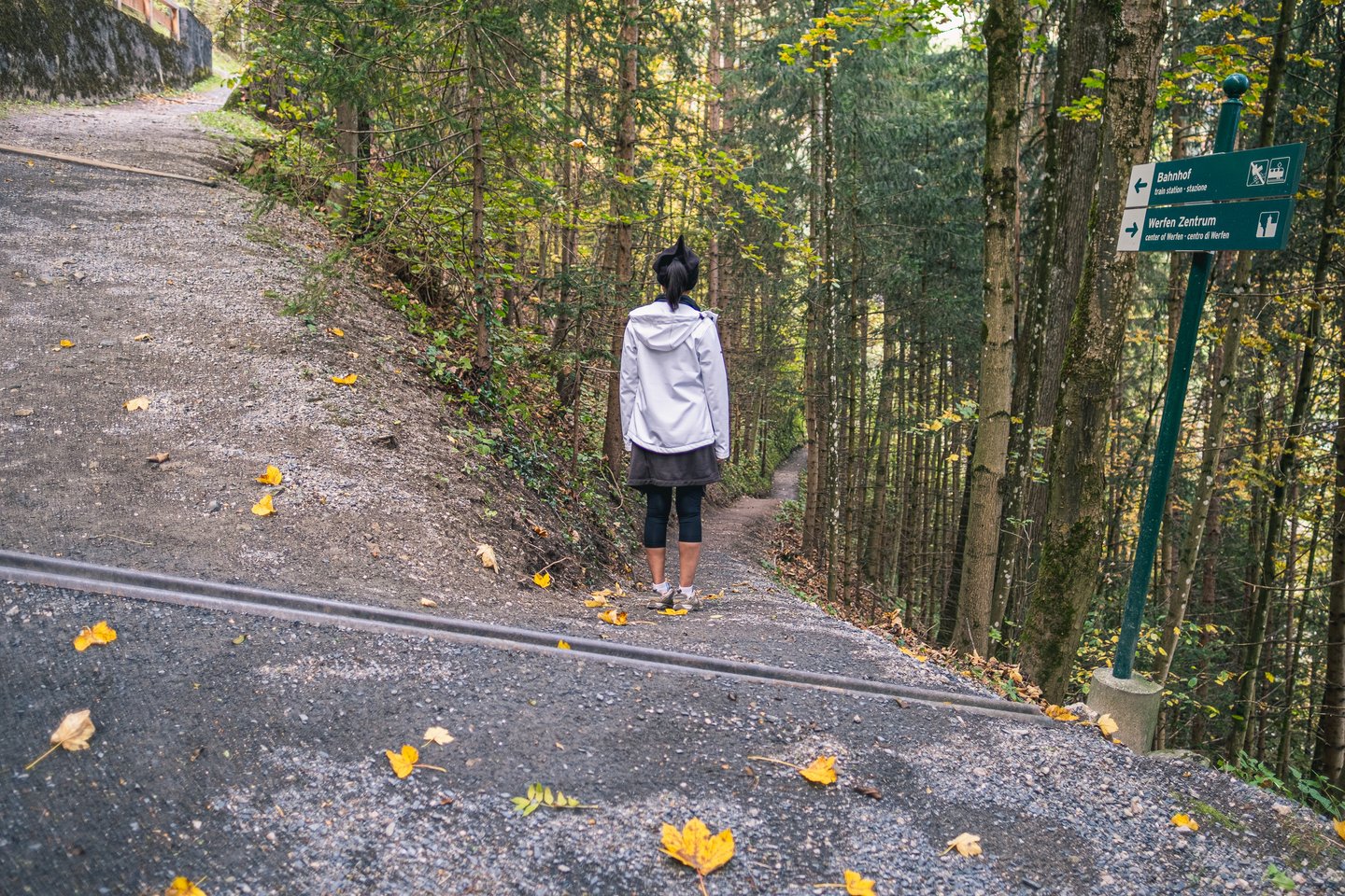 A woman in a white rain jacket stands where a narrow forest footpath leaves the gravel road above Werfen, beside a green sign pointing to the train station and the village centre, with fallen yellow leaves on the track.