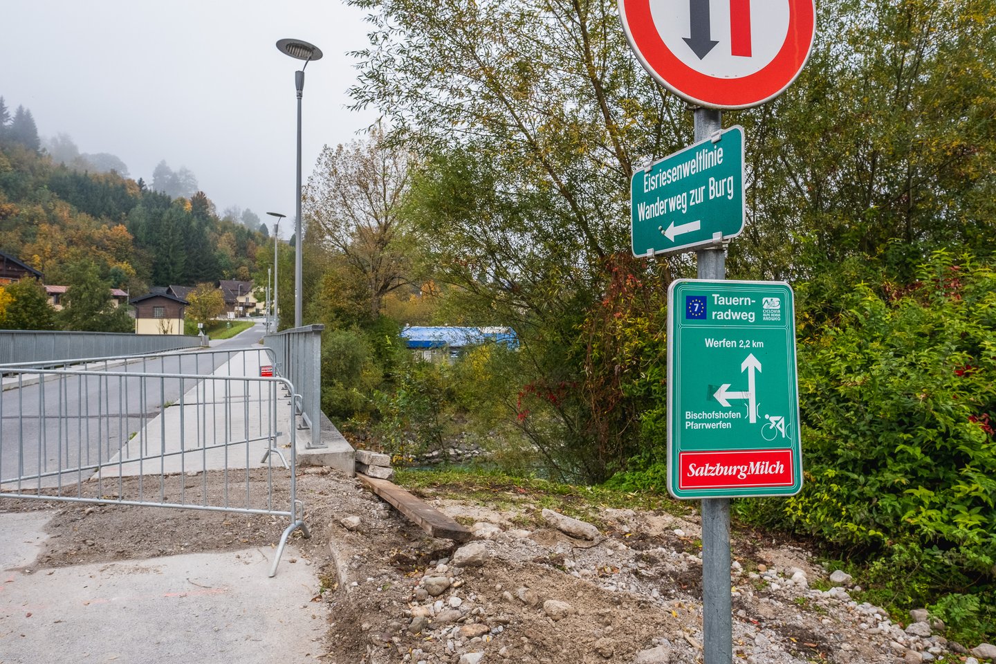 Green direction signs at the end of a river bridge in Werfen, one pointing left to the Eisriesenwelt bus line and the footpath to the fortress, the other marking the Tauernradweg cycle route with Werfen 2,2 km.