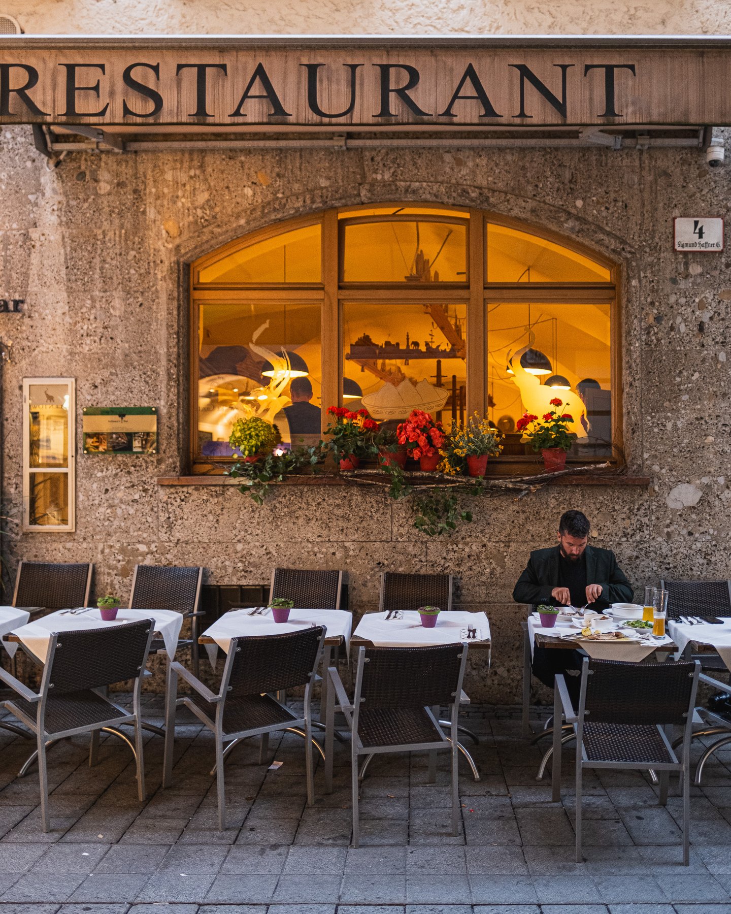 A vertical view of the Wirtshaus Elefant frontage centred on the arched window with its red geraniums and glowing interior, the Restaurant lettering above and the lone diner still eating at the terrace tables below.