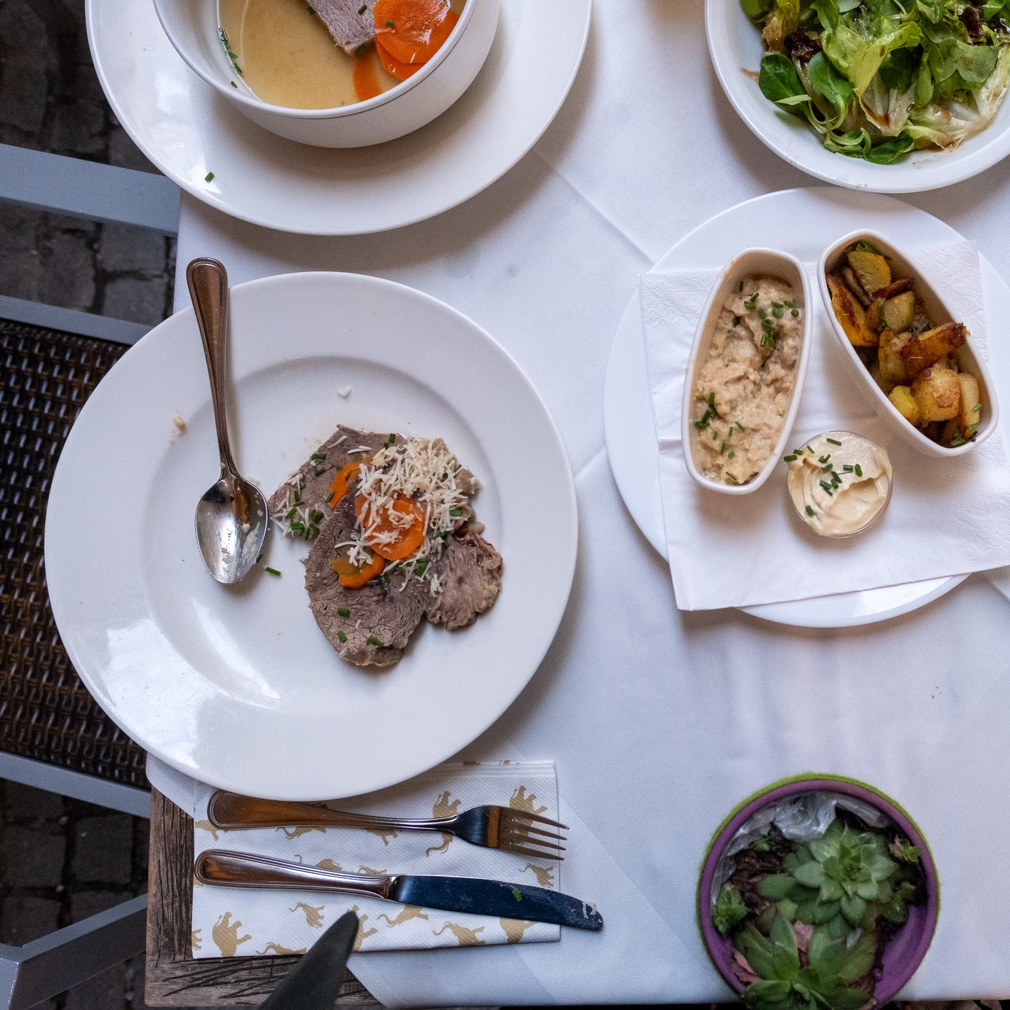 Looking straight down on a terrace table at Wirtshaus Elefant: a bowl of Tafelspitz in broth, a plate of sliced boiled beef with grated horseradish and carrot, roast potatoes, apple horseradish and a green salad on white linen.