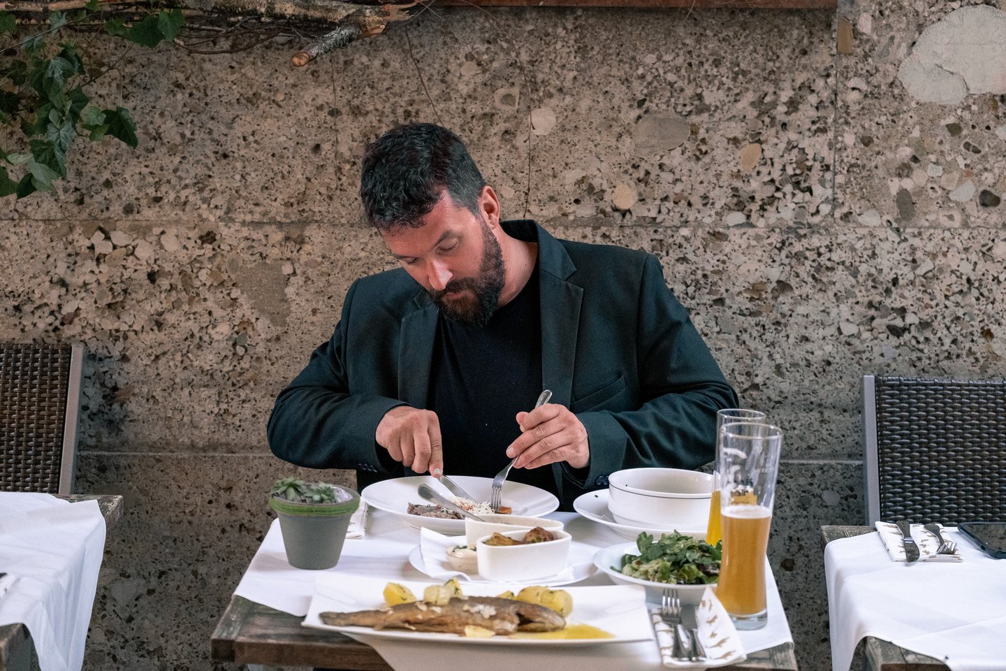 A bearded man in a dark jacket eats Tafelspitz at a white-clothed terrace table set against the pebbly conglomerate wall of the Hotel Elefant building, with a trout, a salad and a wheat beer on the table.