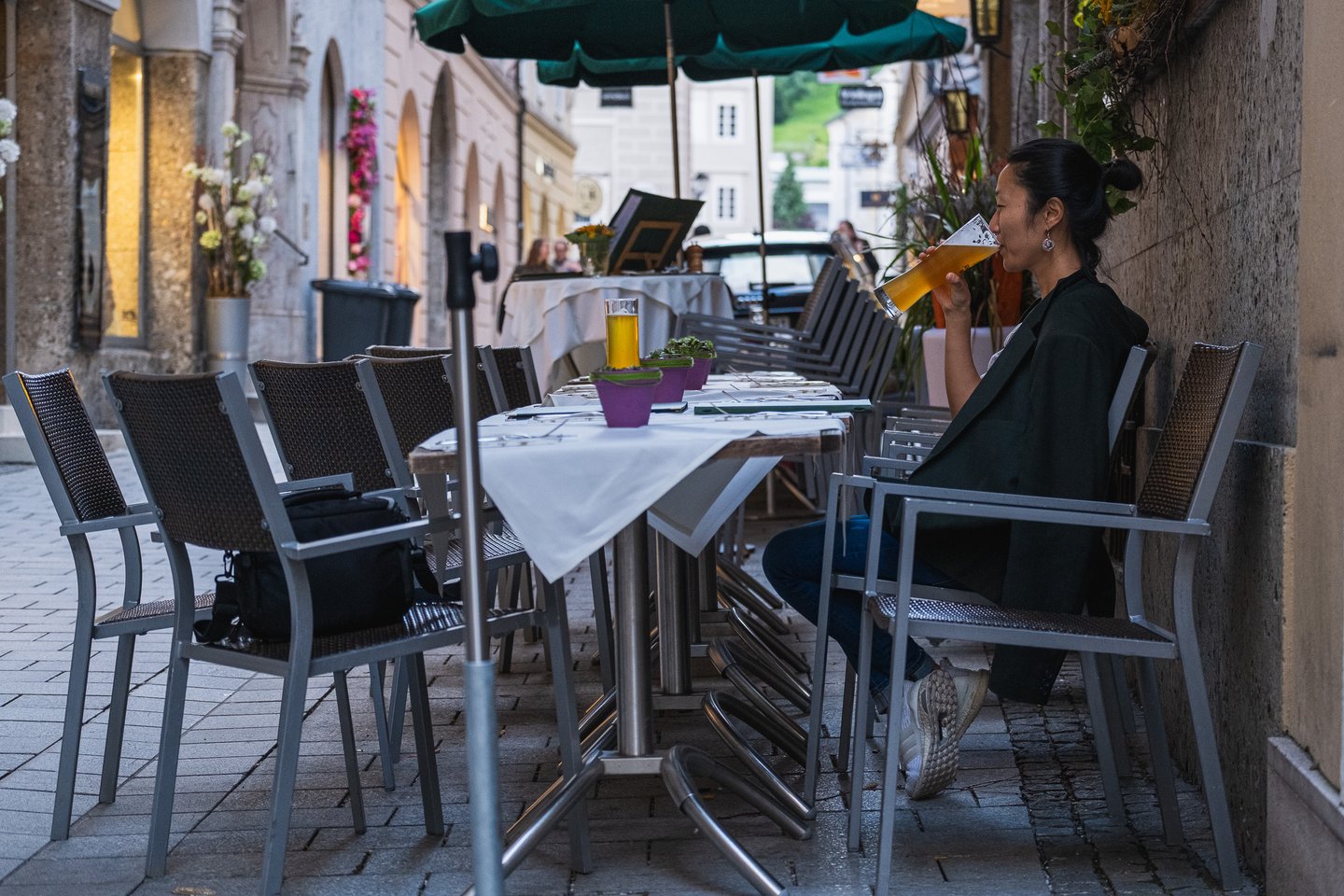 A woman sits alone against the rough stone wall of the lane outside Wirtshaus Elefant and drinks a tall wheat beer, with a row of empty white-clothed tables and grey chairs stretching away under a green parasol.