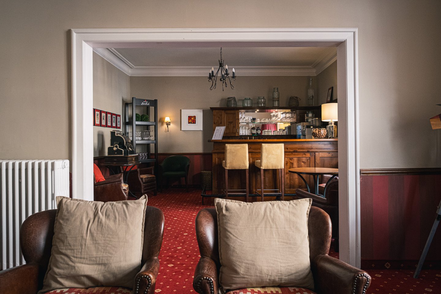 Looking over two leather club chairs through a wide doorway into a small hotel bar in Salzburg, with a wooden counter, pale bar stools, a wrought-iron chandelier and a patterned red carpet.