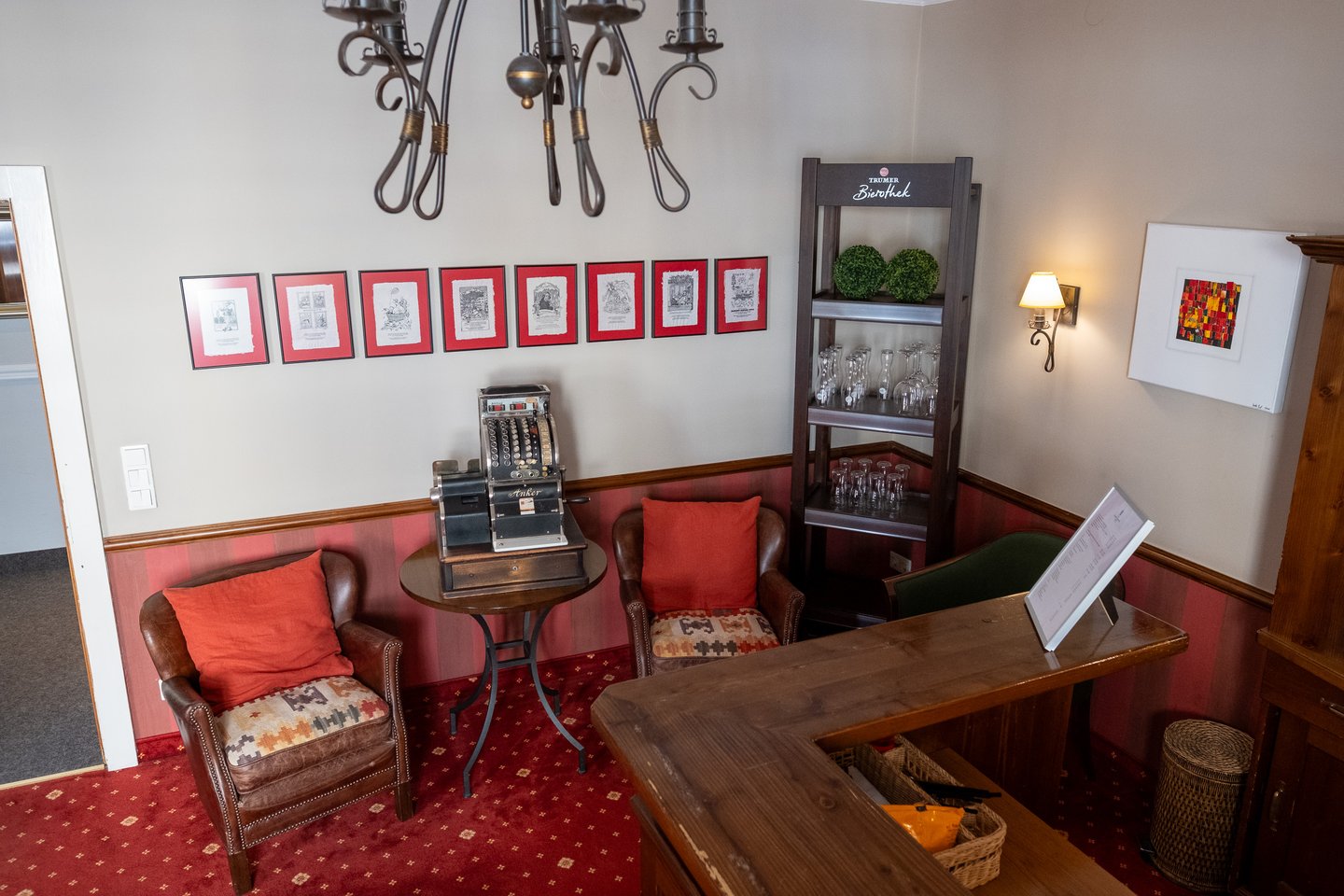 The reception corner of a Salzburg hotel bar, with an antique Anker cash register on a bistro table between two leather armchairs, a row of red-mounted Salzburg prints and a Trumer Bierothek glass shelf.