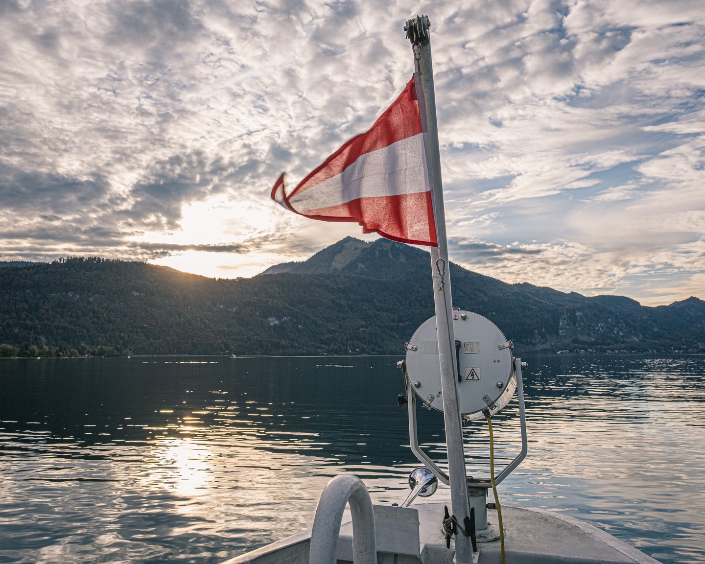 An Austrian flag flies from the bow mast of a Wolfgangsee ferry as the low sun drops behind the wooded ridges across the still lake.
