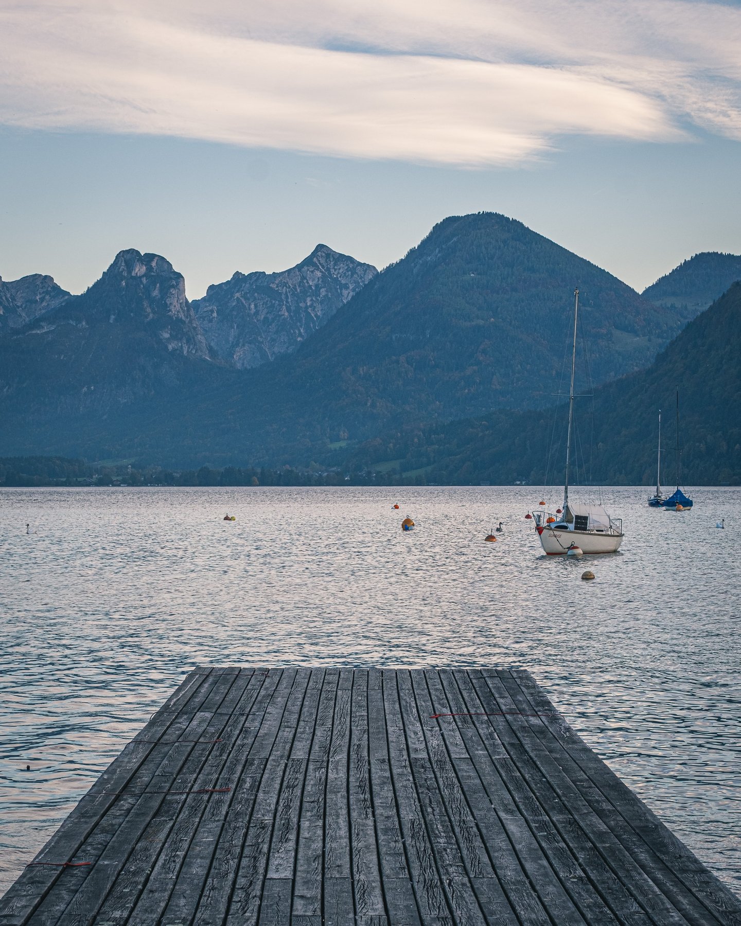 A broad wooden jetty runs out into the still Wolfgangsee at dusk, with a white sailing boat and two others moored to buoys and rocky peaks silhouetted beyond the far shore.