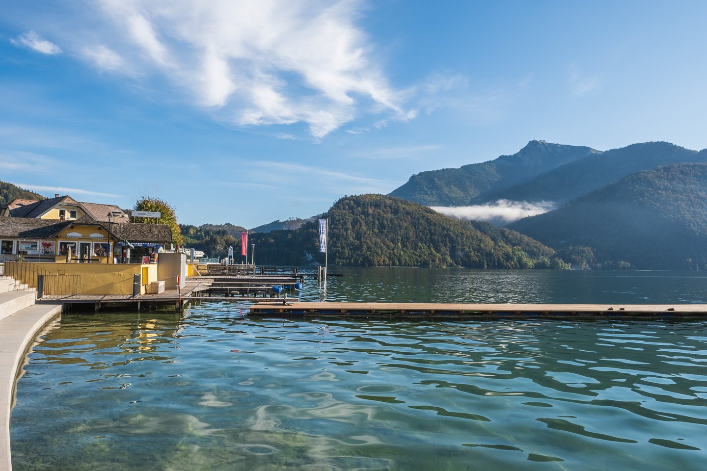 Wolfgangsee lies glassy in low morning sun, seen past the St. Gilgen landing stage, with a band of cloud caught on a wooded ridge below a rocky summit.