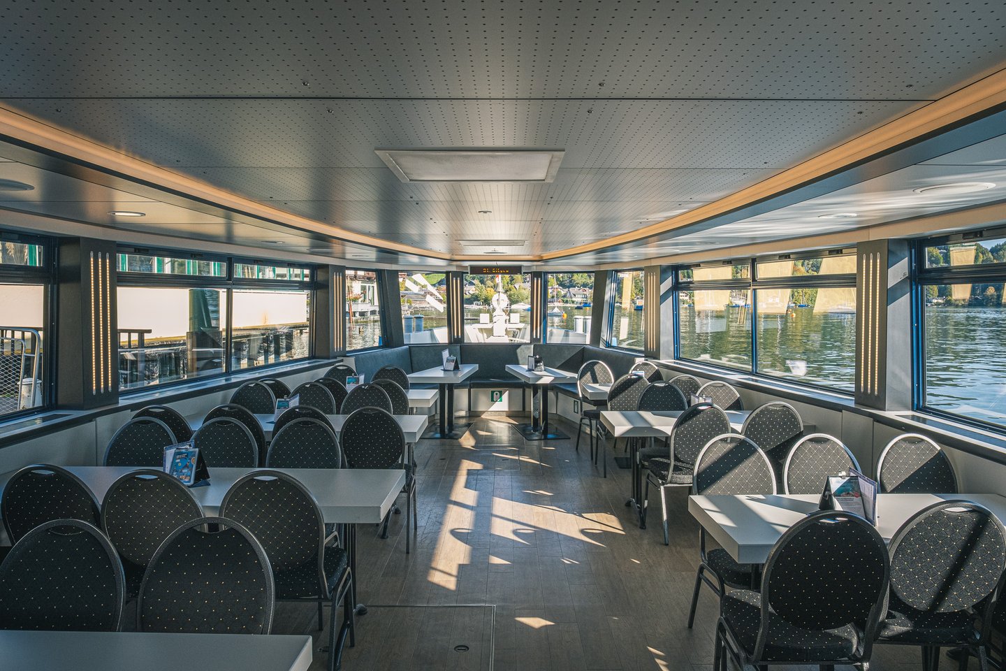 The empty passenger saloon of a Wolfgangsee ferry, rows of tables and dark upholstered chairs with the lake and moored boats through the windows.