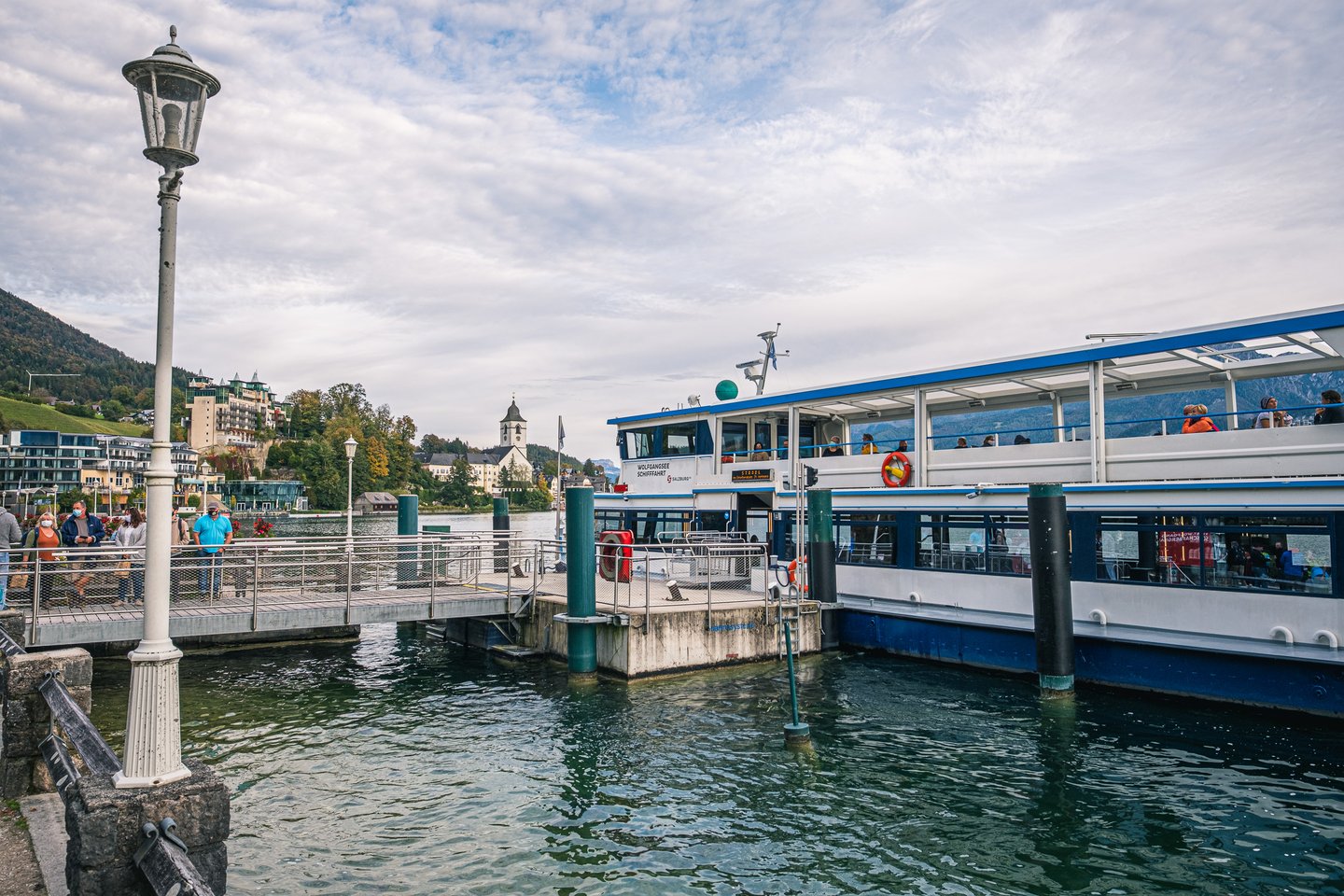 A Wolfgangsee Schifffahrt passenger ferry lies at the St. Wolfgang landing stage while masked passengers wait on the jetty, the village church tower rising across the water.
