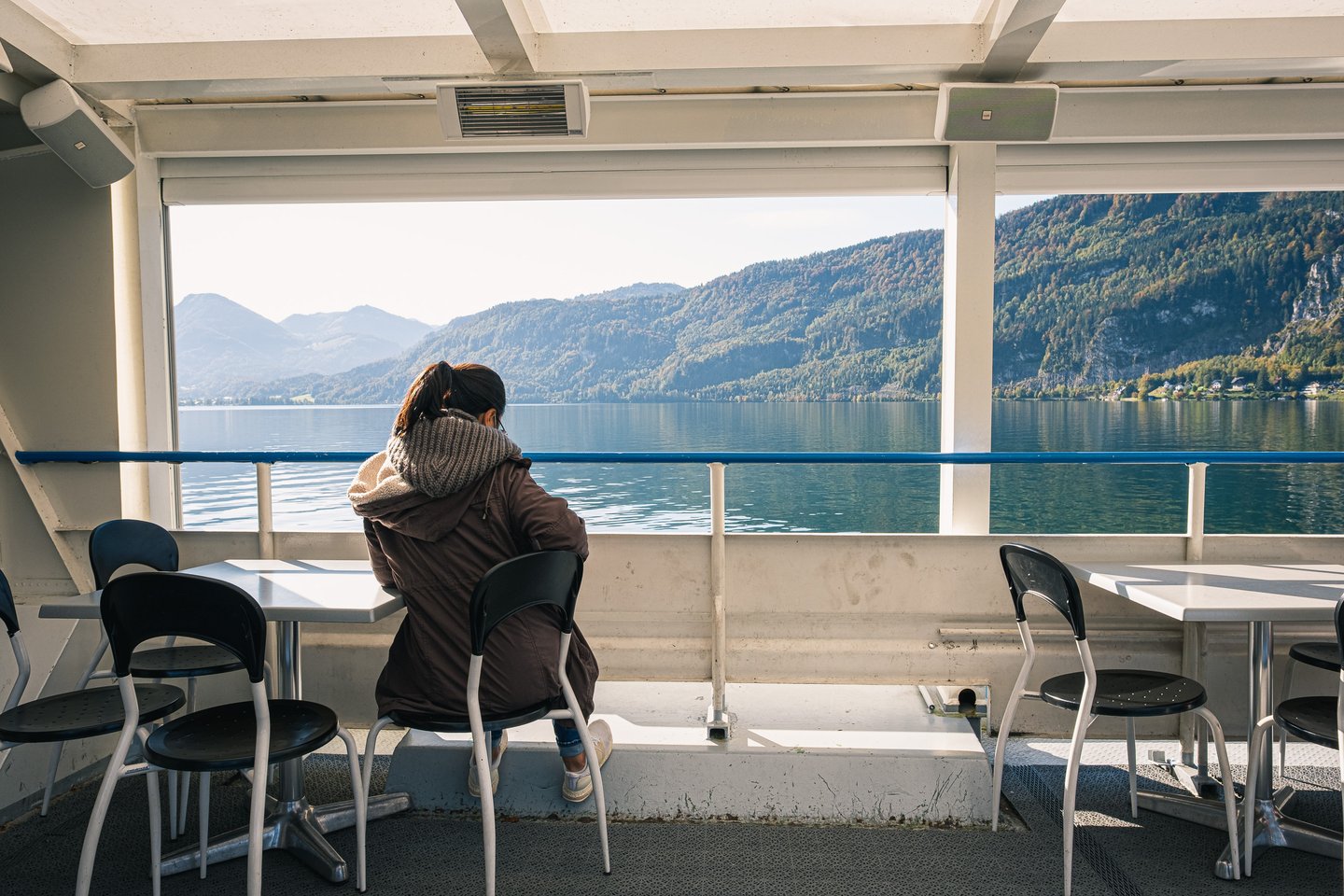 A single passenger in a winter coat and scarf sits at a deck table on a Wolfgangsee ferry, looking out over the still lake and its autumn-coloured slopes.