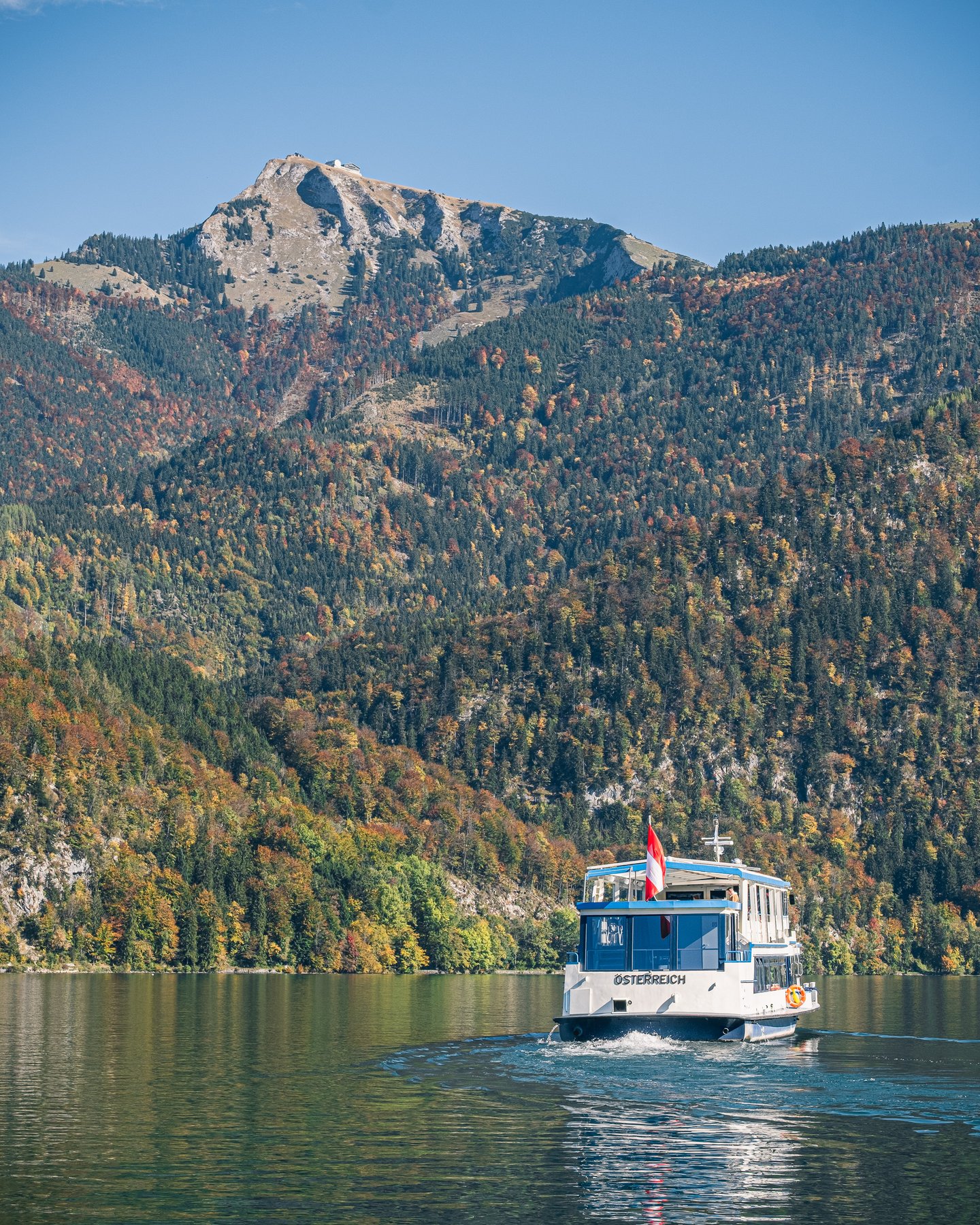 The ferry MS Österreich crosses the Wolfgangsee below the Schafberg, whose bare rock summit rises above slopes of red and gold autumn forest.