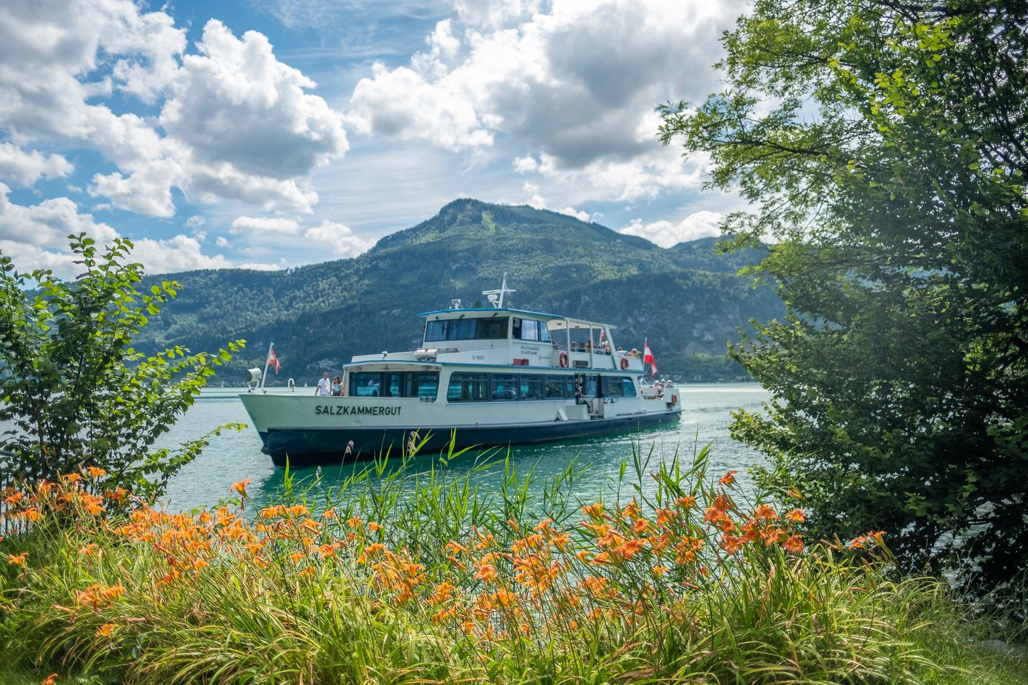 The Wolfgangsee ferry MS Salzkammergut crosses the turquoise lake, framed by a bank of orange daylilies in the foreground.