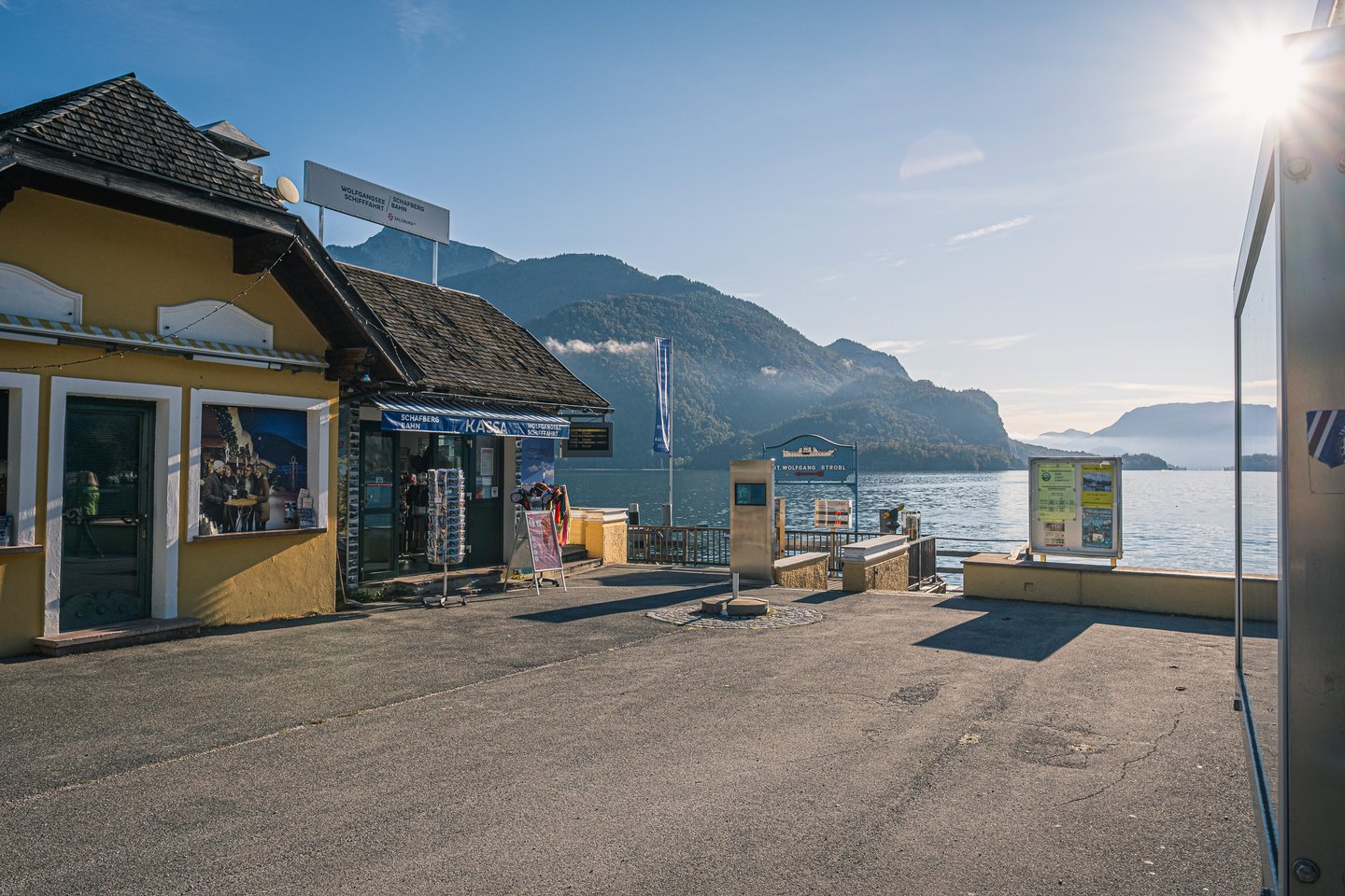 The yellow ticket house at the St Gilgen pier, signed Wolfgangsee Schifffahrt and Schafbergbahn above a Kassa window with a postcard stand outside, stands empty in low morning light with the lake and misted ridges beyond.