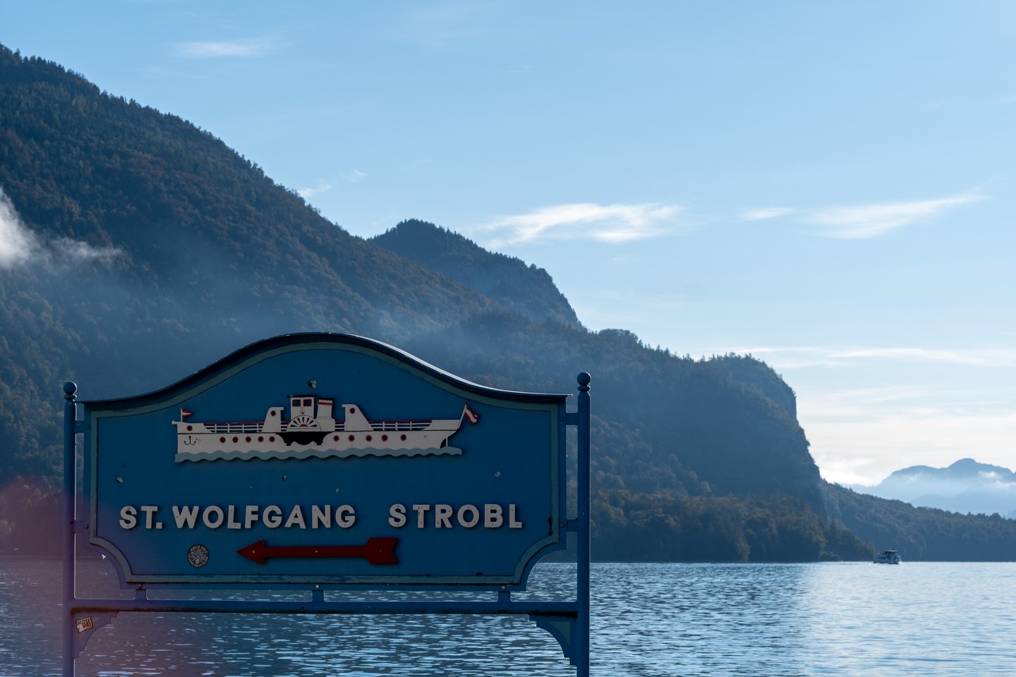 A blue landing-stage board carrying a white relief paddle steamer and the words St. Wolfgang Strobl with a red arrow points left across a misty autumn Wolfgangsee, a boat small in the distance.