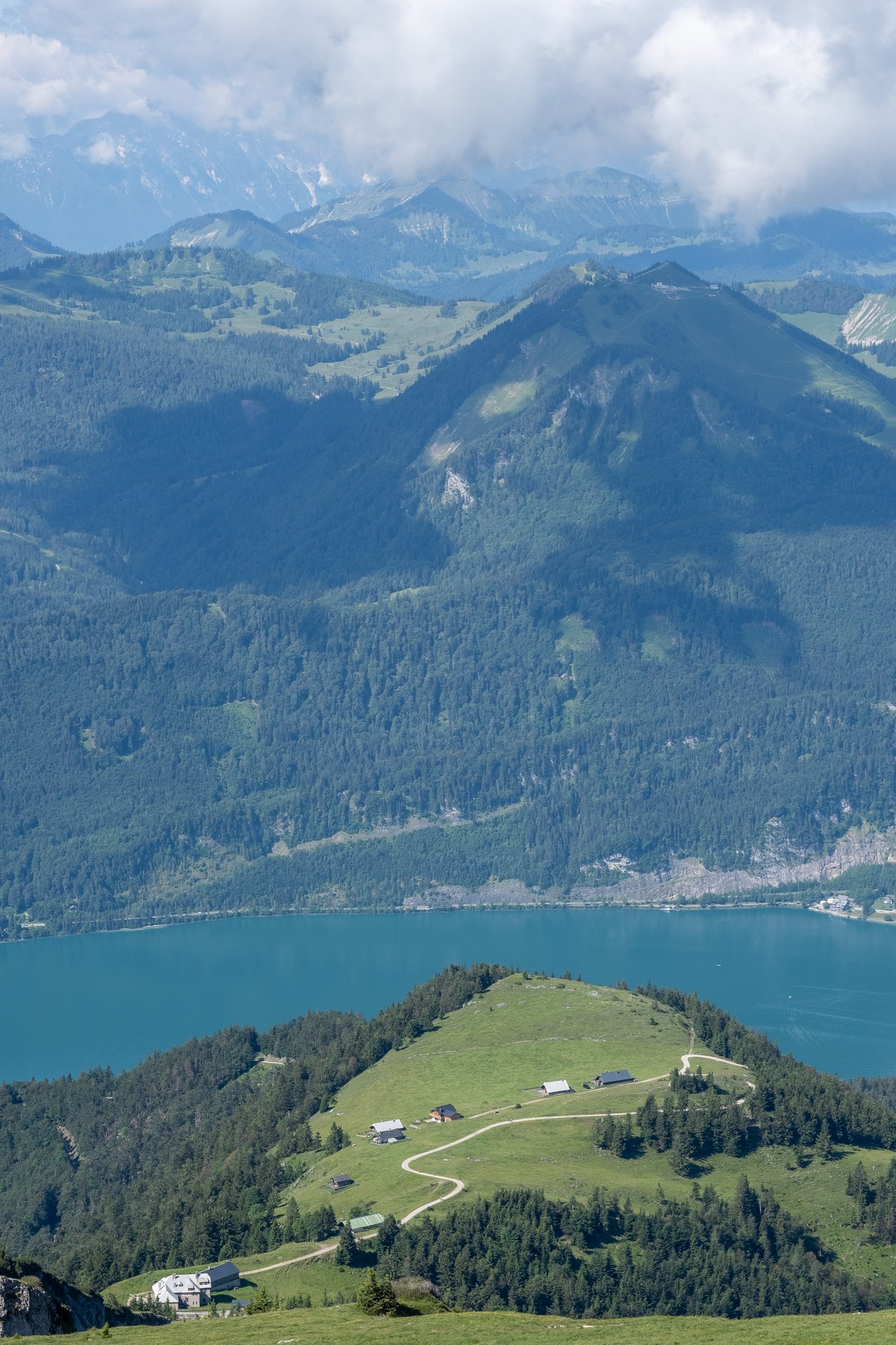 Looking from the Schafberg over a grassy spur dotted with alm huts to the deep blue Wolfgangsee, with a summit carrying buildings beyond it and cloud capped limestone ranges on the horizon.