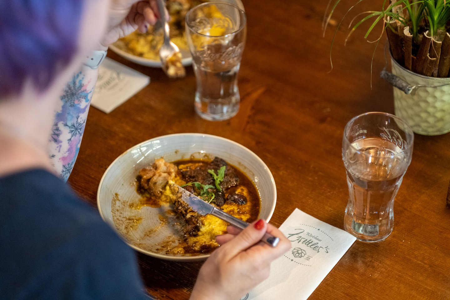 A woman cuts into a bowl of braised beef with polenta and rocket at Zwettler's Wirtshaus in Salzburg, a napkin printed with the tavern's hop logo lying beside the bowl.