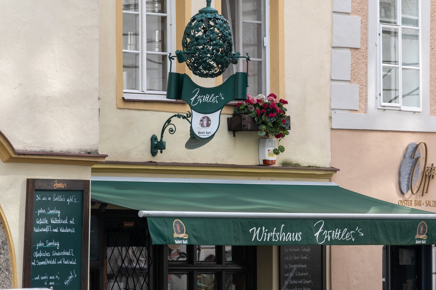 A green wrought-iron guild sign lettered Zwettler's hangs beside a geranium box above the green Wirtshaus Zwettler's awning in Salzburg, with a handwritten menu board at the left.