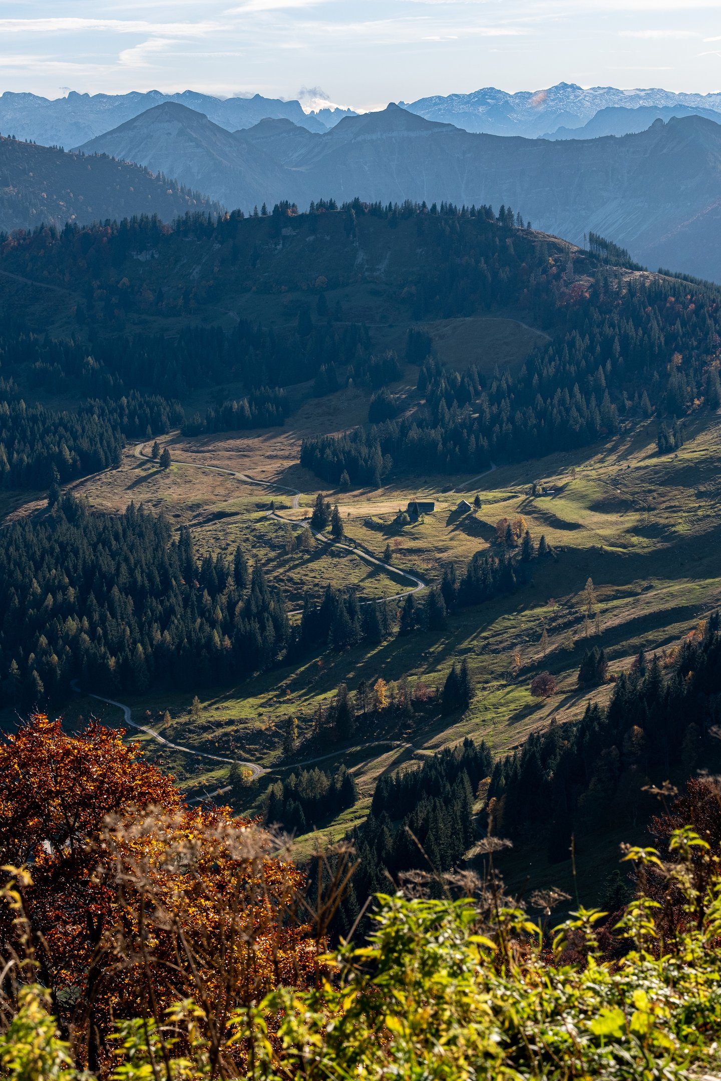 Alpine pastures with a handful of dark timber huts and a pale gravel road curve below the Zwölferhorn, with hazy, snow-dusted ranges on the far horizon.