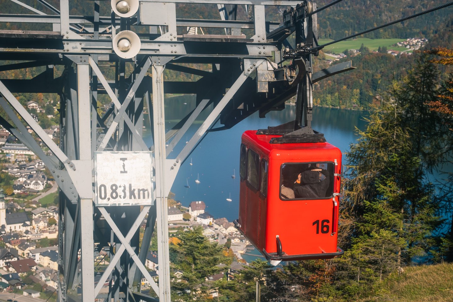 A red cable car cabin numbered 16 passes a lattice pylon marked 0,83 km, high above the roofs of St. Gilgen and the blue Wolfgangsee.