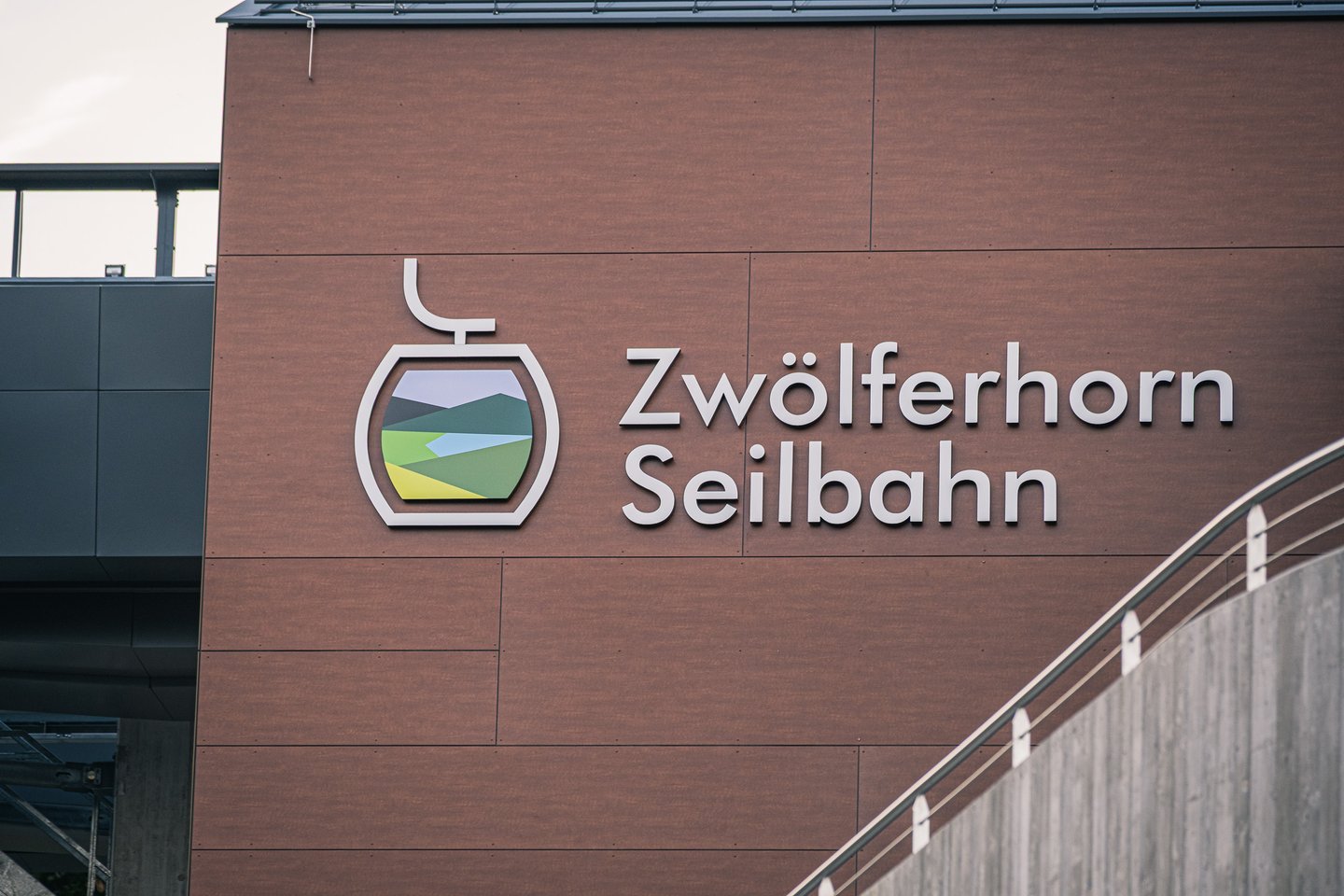 The words Zwoelferhorn Seilbahn and a gondola-shaped logo showing green slopes and a blue lake stand in white relief on the red-brown cladding of the valley station.
