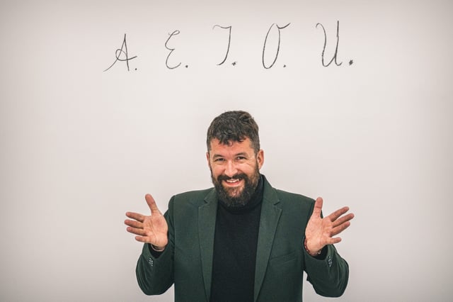 A smiling bearded man in a dark green blazer and black turtleneck spreads both hands wide, standing beneath the letters A. E. I. O. U. written in cursive on a pale wall.