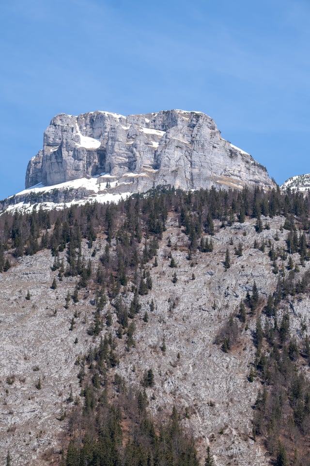 A broad blocky limestone summit wall above Altaussee, streaked with late spring snow and pierced by a dark natural rock window, rises above scree slopes and scattered spruce.