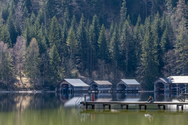 A row of wooden boathouses on stilts lines the far shore of the Altausseer See below a spruce forest, with a woman sitting alone on the long jetty in the foreground.