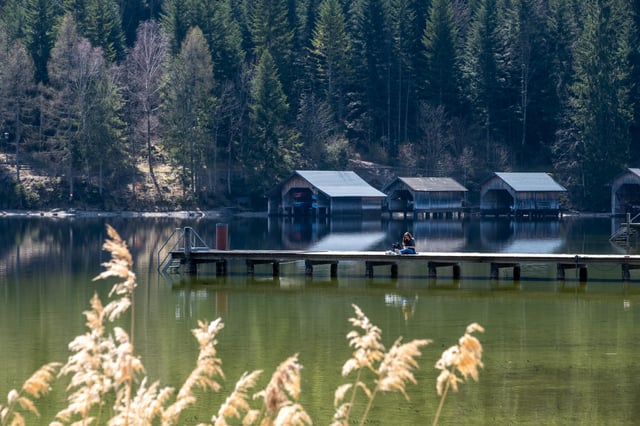 Dry reed plumes in the foreground frame a row of wooden boathouses across the Altausseer See, with a woman sitting on a long jetty in the middle distance.