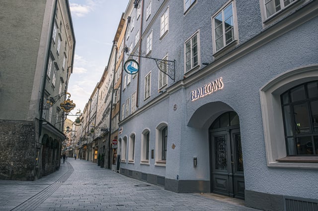 Looking steeply up the blue-grey facade of the Arthotel Blaue Gans on an almost empty Getreidegasse, with the goose guild sign and the lit BLAUEGANS lettering above the dark arched door.