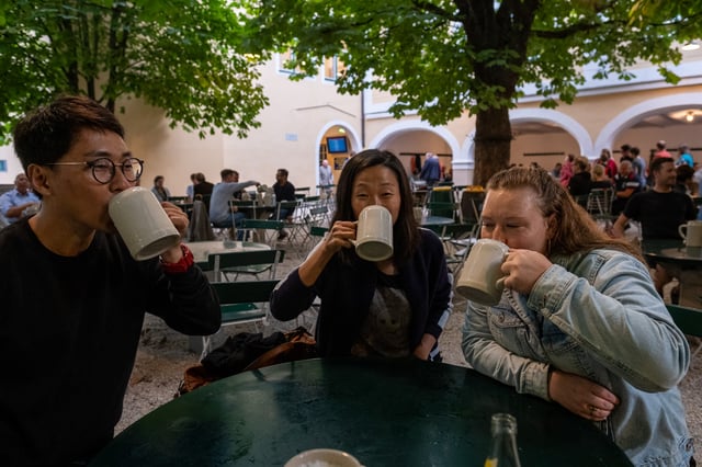 Three friends drink at the same moment from heavy stoneware mugs at a green table under the chestnut trees of the Augustiner Braustubl beer garden in Mulln.