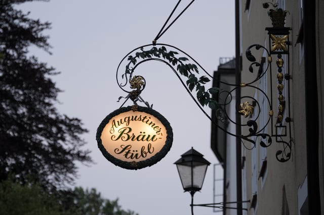 The illuminated oval sign of the Augustiner Braeustuebl hangs from an ornate wrought-iron bracket wound with hop leaves against a dusk sky in Salzburg.
