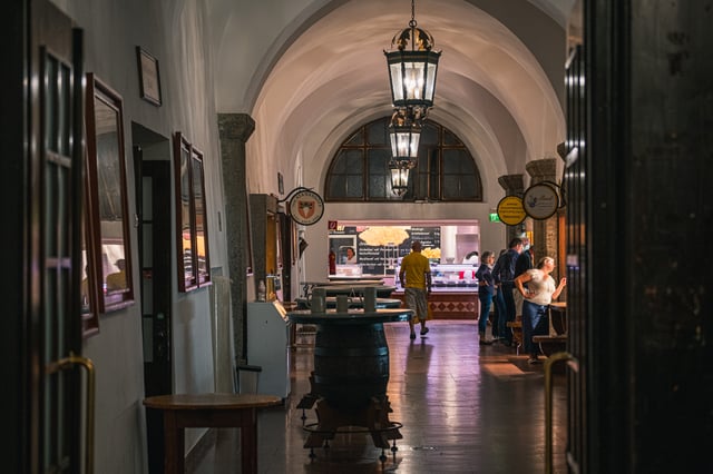 Lanterns hang the length of the white vaulted food arcade of the Augustiner Braustubl, past barrel tables and hanging stall signs to a lit counter where guests wait at the far end.