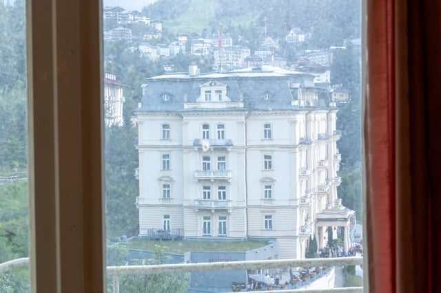 A white belle epoque hotel with a slate mansard roof seen through a hotel window pane in Bad Gastein, the town climbing the wooded slope behind it.