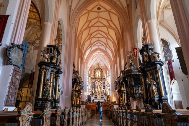 A visitor stands alone in the central aisle of the Mondsee basilica looking up, with carved pew ends in the foreground and the high altar lit at the far end.