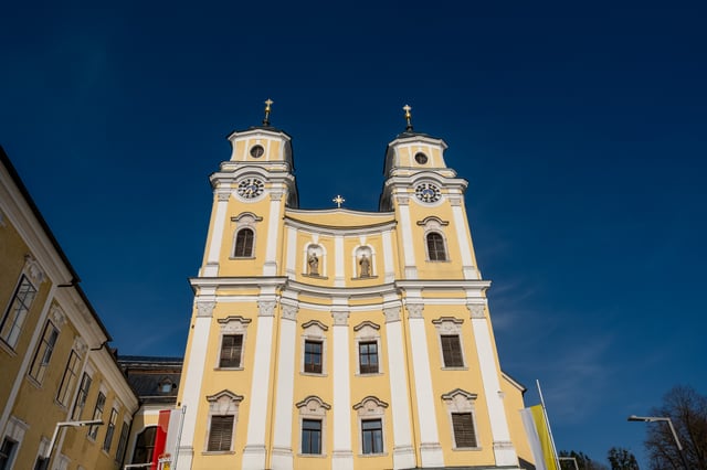 The yellow and white twin tower facade of the Mondsee basilica photographed steeply from below against a deep blue sky, with gold crosses on both onion cupolas.