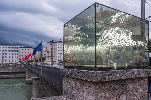 Glass cube of the Beyond Recall installation with white cursive neon script on the Staatsbruecke over the Salzach in Salzburg