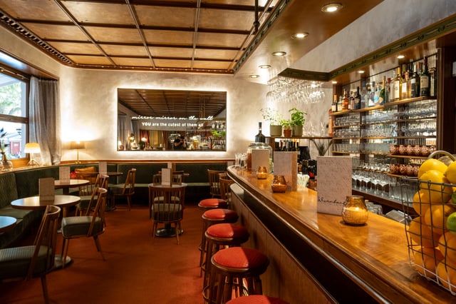 The polished wooden bar of Cafe Wernbacher runs to the right with red-topped stools, backlit shelves of glassware and spirits, herb pots and a wire basket of lemons, the green banquettes and lettered mirror to the left.