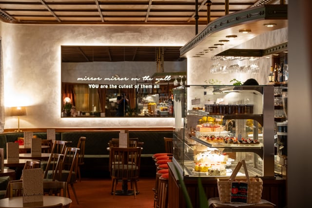 A lit glass cake cabinet of tarts, cream slices and fruit pastries stands at the end of the Cafe Wernbacher bar, facing the green banquettes and the lettered mirror across the red carpet.