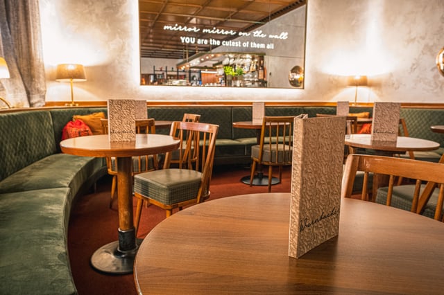 A textured Wernbacher menu folder stands upright on a round wooden table in the foreground, with the lettered mirror, green banquettes and spindle-back chairs of the cafe behind it.