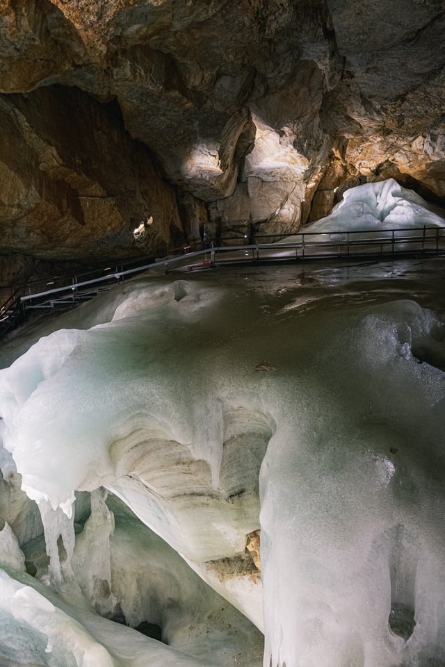 A frozen cascade of milky green ice with a fringe of icicles fills the foreground of the cave, the visitor walkway a thin line along the rock wall behind it.