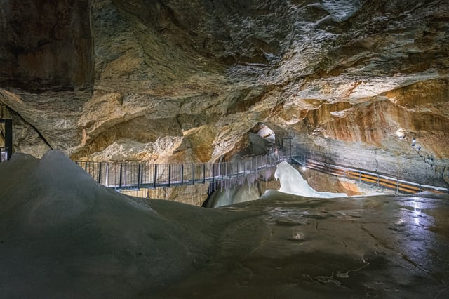 A long steel walkway bridge crosses high above the grey ice floor of a broad chamber in the Dachstein Giant Ice Cave, with icicles hanging from its underside.