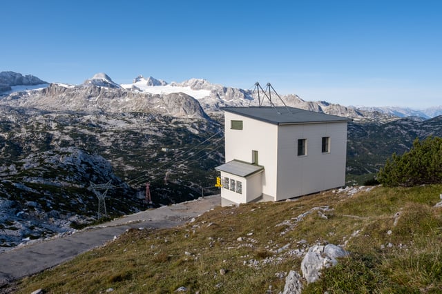 A small white cable car station building sits on the grassy edge of the Krippenstein plateau above Obertraun, its haul rope dropping away past a red lattice pylon while the Dachstein massif and its glacier fill the horizon.