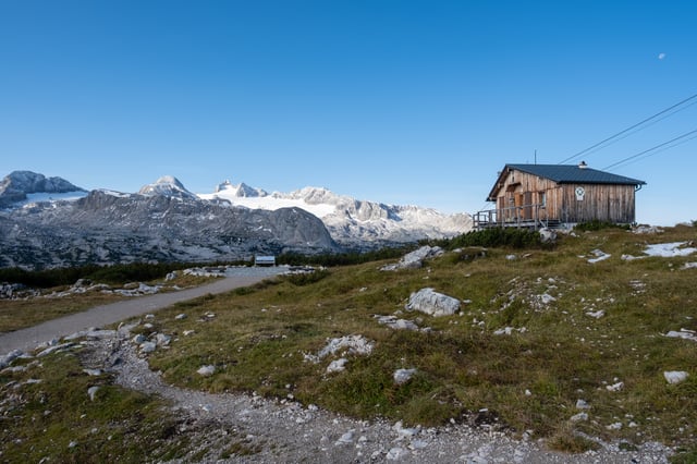 A small weathered wooden hut sits on the grassy Krippenstein plateau in early morning light, facing the long snow-topped wall of the Dachstein range with a pale moon overhead.