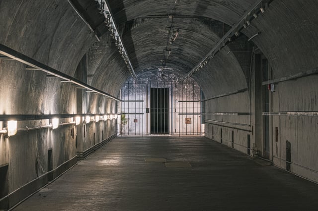A long barrel vaulted concrete corridor in the Obersalzberg bunker, lit by bulkhead lamps along the left wall and closed at the end by a floor to ceiling steel grille in front of a doorway, the walls around it covered in scratched visitor graffiti.