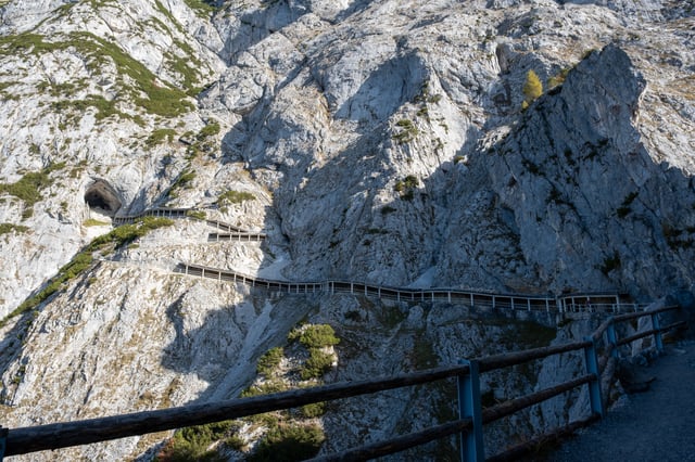 The long railed walkway traverses the limestone cliff and climbs in stages towards the dark mouth of the Eisriesenwelt at the left, with the path's own timber and steel railing crossing the foreground.