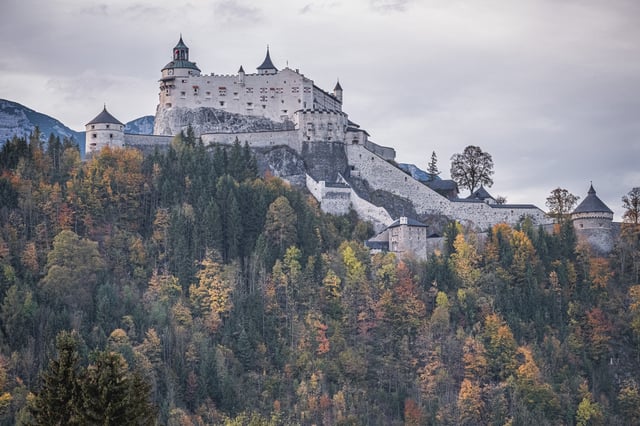 Festung Hohenwerfen sits white on its rock above a hillside of orange and green autumn forest, its keep, clock tower and ring of round bastion towers set against a flat grey sky.